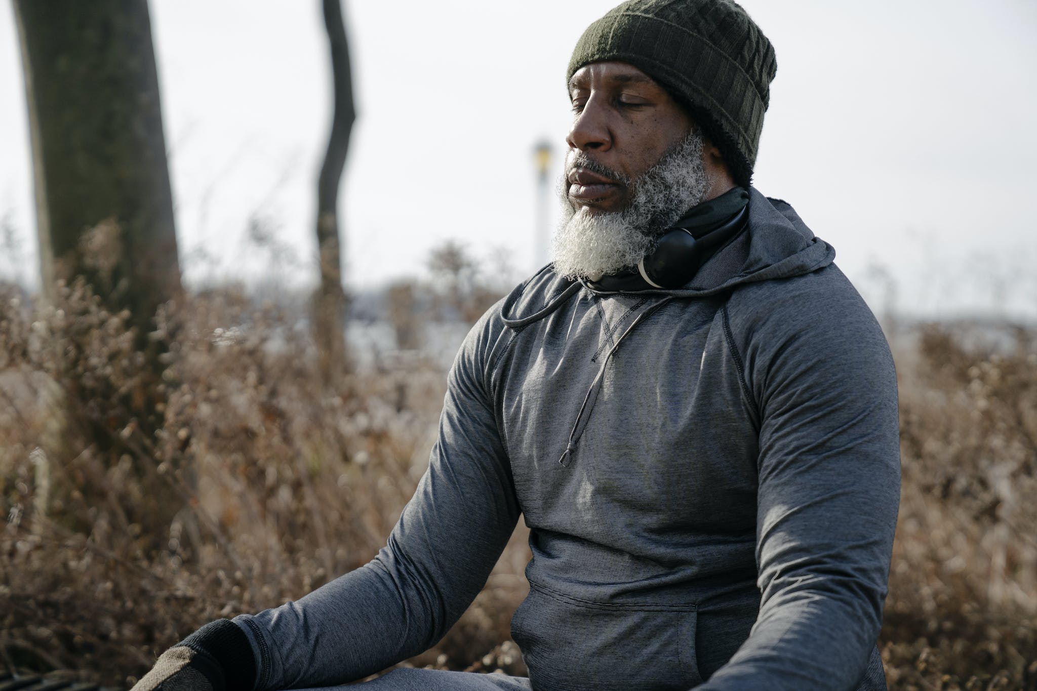 Bearded African American male in warm activewear and knitted hat sitting and meditating with closed eyes in autumn nature