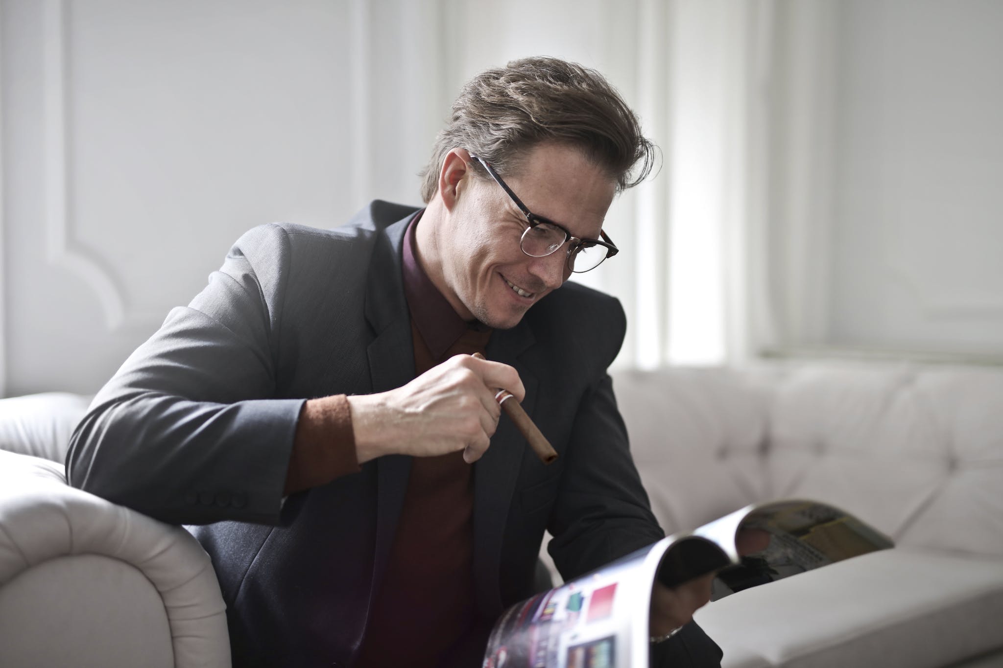 Smiling rich gentleman wearing classy suit and stylish eyeglasses sitting with cigar on leather sofa in luxury living room and reading magazine